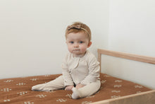 Load image into Gallery viewer, baby girl sitting up wearing organic cotton vanilla color footed zipper pajamas and a bow head band