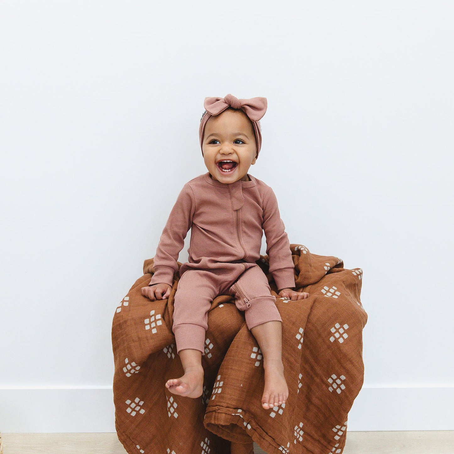 smiling baby girl wearing matching organic cotton footless zipper pajamas and head wrap bow by mebie baby dusty rose color
