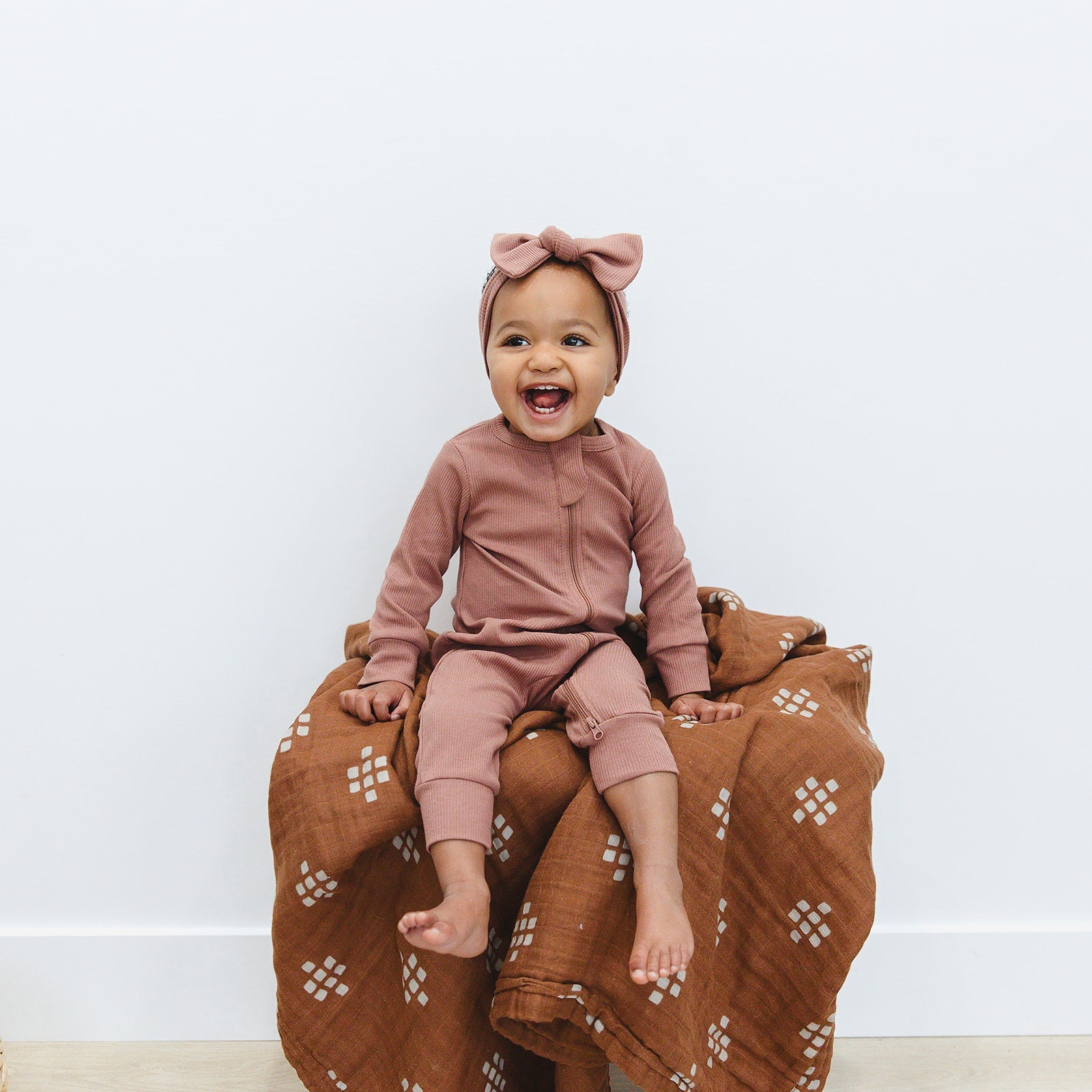 smiling baby girl wearing matching organic cotton footless zipper pajamas and head wrap bow by mebie baby dusty rose color